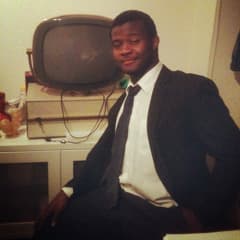 Young man in a suit and tie sitting in a chair during an indoor speech practice session.