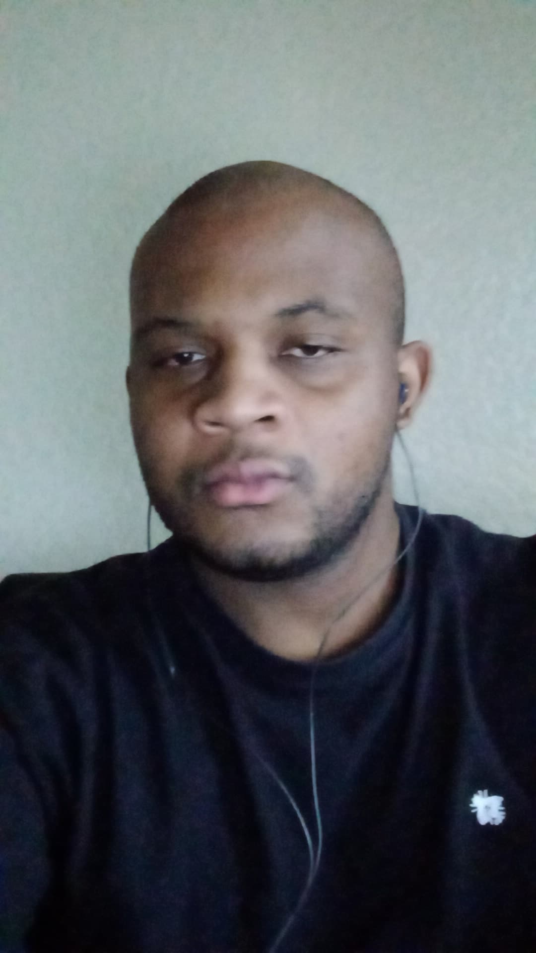 Close-up selfie of a bald man wearing a black t-shirt and wired earbuds against a plain wall.