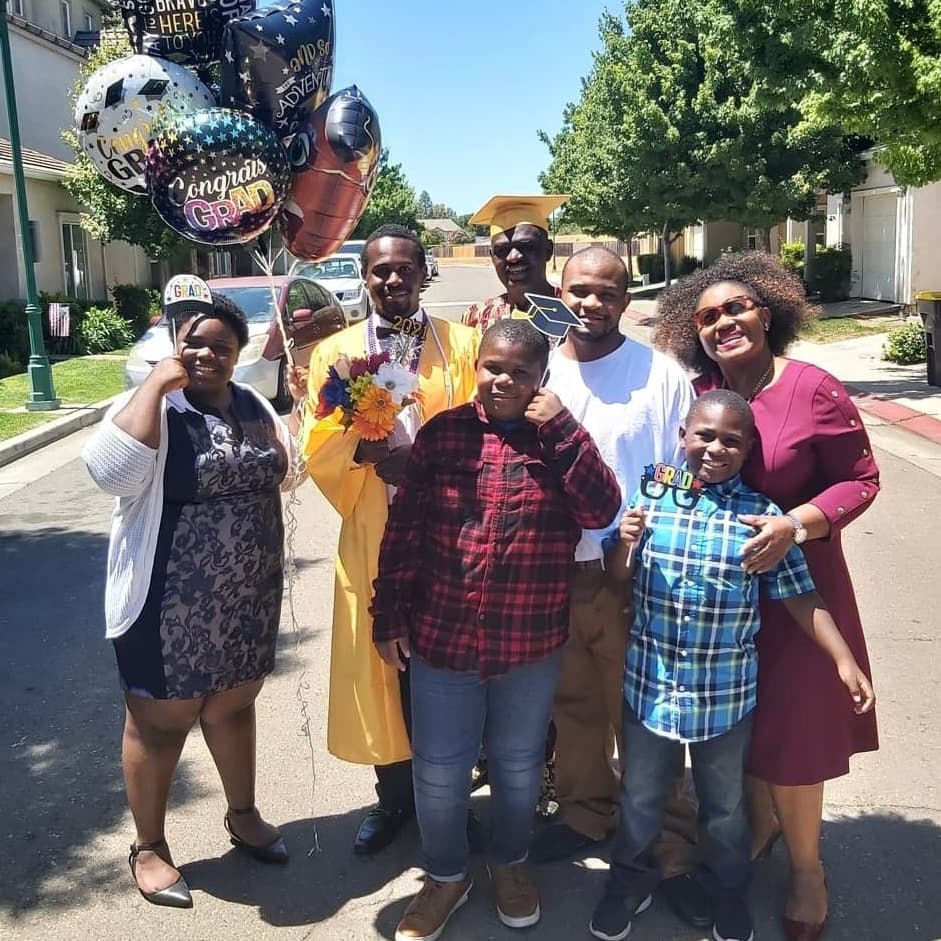 A family posing together outdoors celebrating a 2021 graduation, with a graduate in cap and gown and balloons reading Congrats Grad.