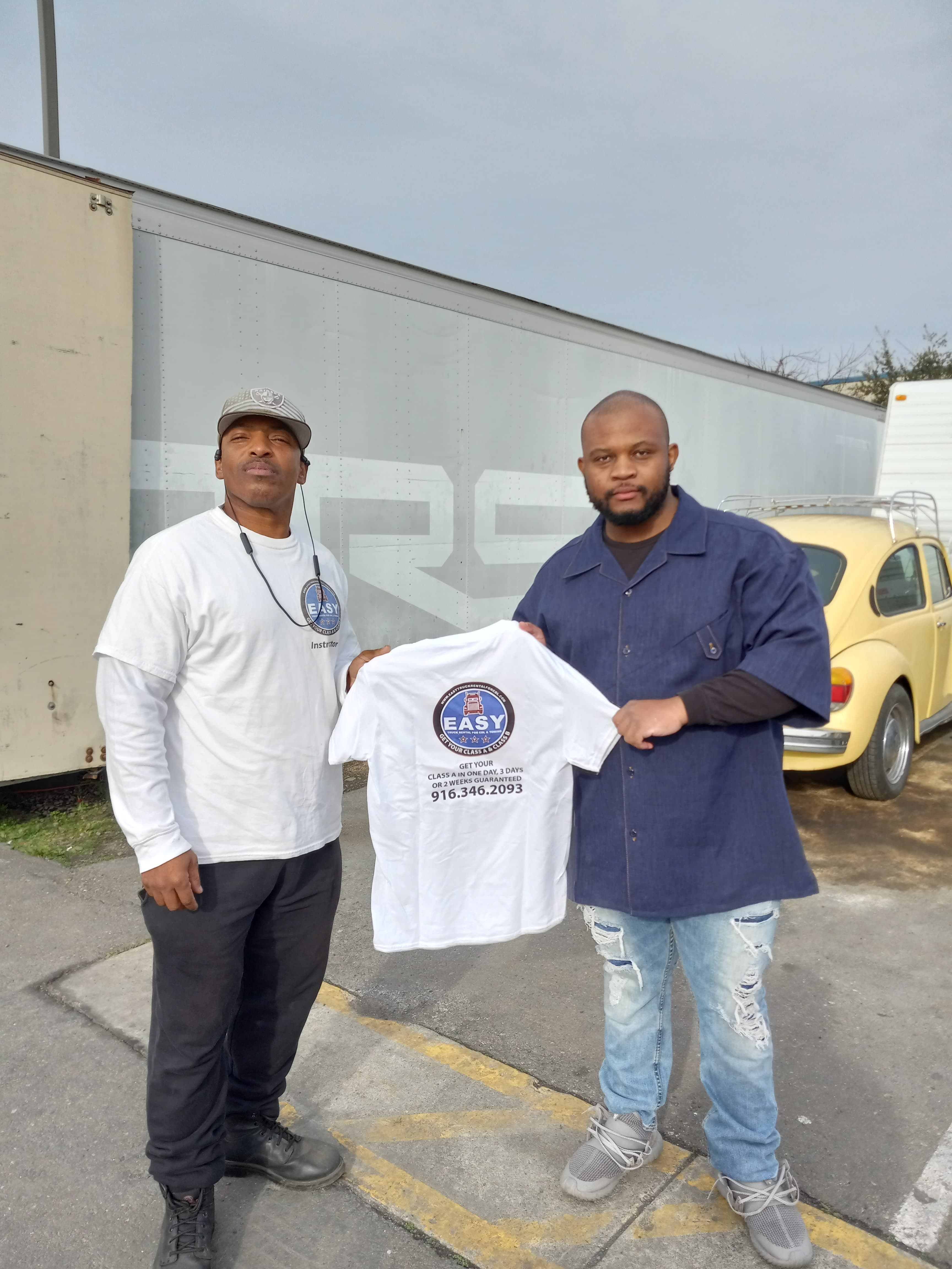 Two men standing in front of a semi-truck trailer, one holding a white t-shirt for a CDL training school, the other wearing a matching shirt and cap labeled Instructor.