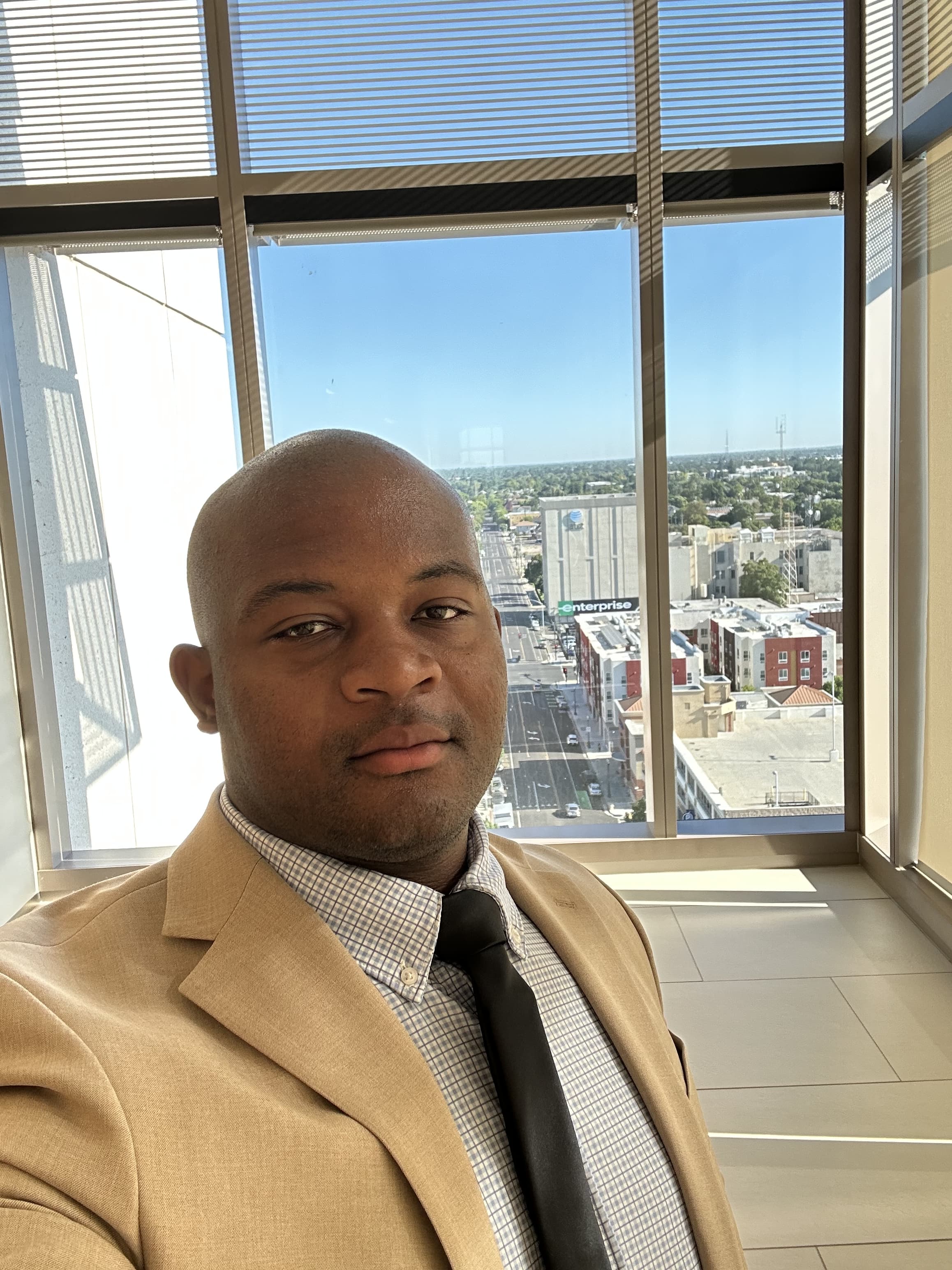 Man in a tan suit and checked shirt smiling in front of a floor-to-ceiling office window overlooking a city street.