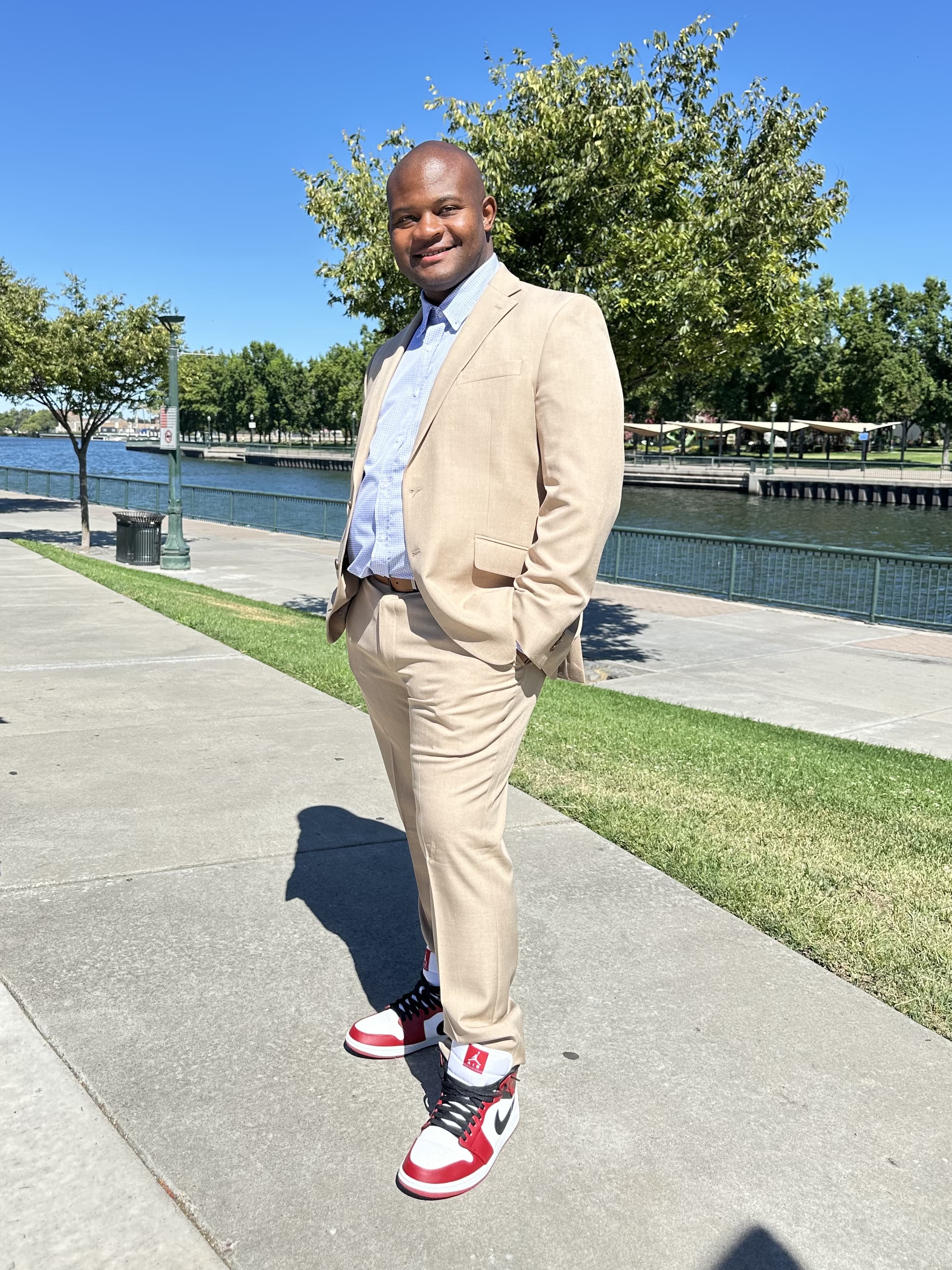 Man in a tan suit and red-and-white sneakers standing on a paved waterfront path with trees and water in the background.
