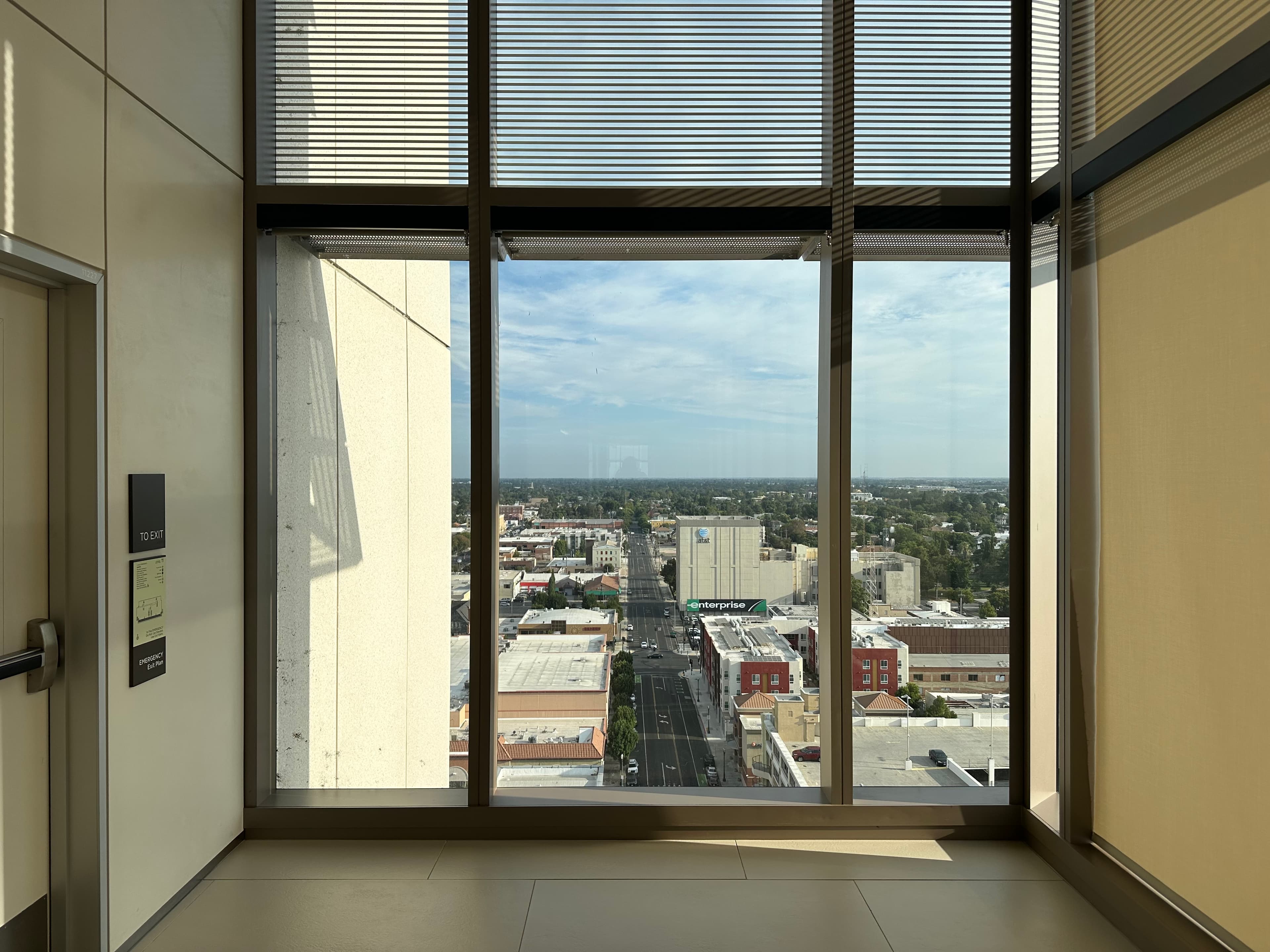 View from inside a modern building through floor-to-ceiling windows onto a sunlit cityscape, road, and trees under a blue sky.
